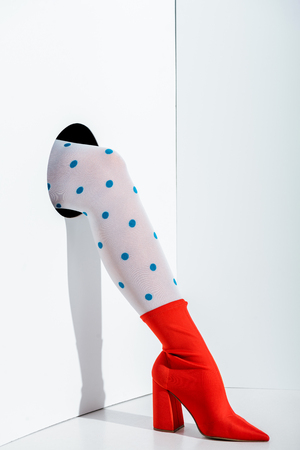 cropped image of girl showing leg in trendy white tights with blue dots and red shoe in hole on whiteの写真素材