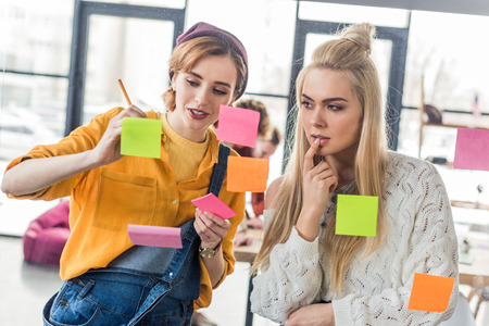 beautiful casual businesswomen looking at colorful sticky notes on glass window in office