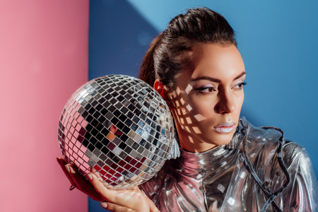 beautiful stylish young woman posing with silver disco ball on pink and blue backgroundの写真素材