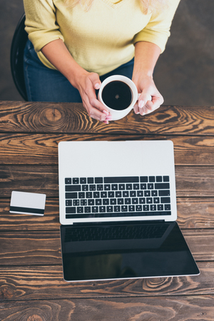 cropped view of woman holding cup of coffee near laptop and credit cardの写真素材