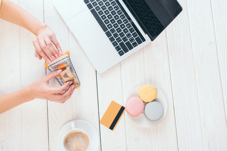 top view woman holding toy shopping trolley with small paper boxes near laptop, cup and macaroons