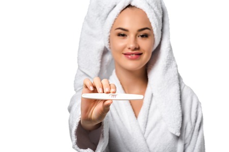 attractive girl in bathrobe smiling, showing pregnancy test and looking at camera isolated on whiteの写真素材