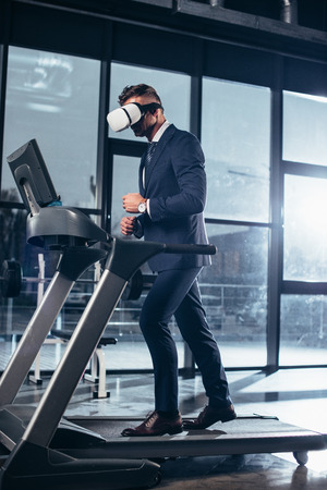 side view of businessman in suit and virtual reality headset exercising on treadmill in gymの写真素材