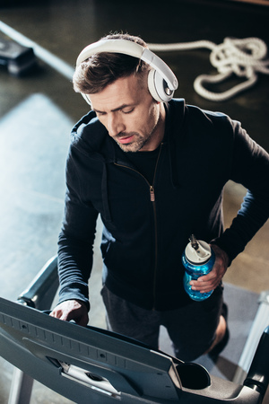 high angle view of handsome sportsman in hoodie exercising on treadmill and holding sport bottle in gymの写真素材