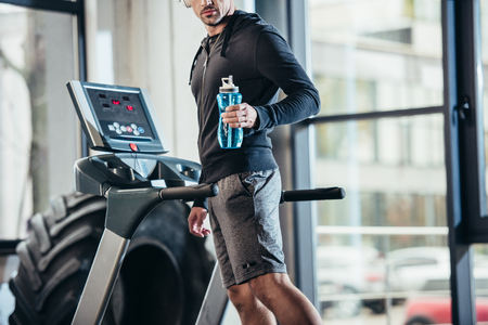 cropped image of sportsman in hoodie exercising on treadmill and holding sport bottle in gymの写真素材