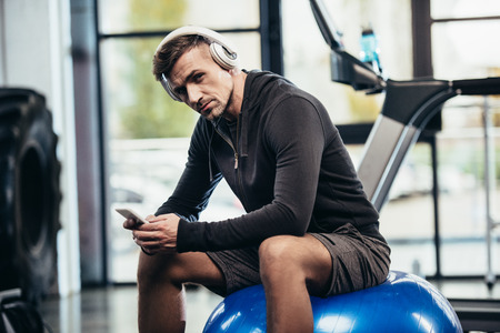 handsome sportsman sitting on fitness ball, holding smartphone and looking at camera in gym