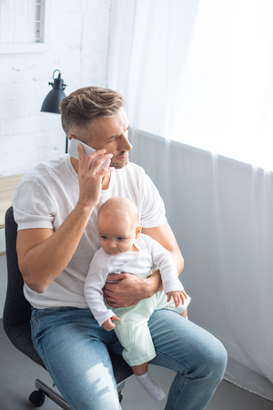 handsome father sitting on chair, talking on smartphone and holding baby daughter at homeの写真素材