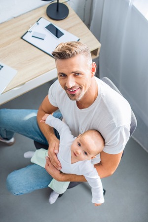 smiling father sitting on chair, holding baby daughter and looking at camera