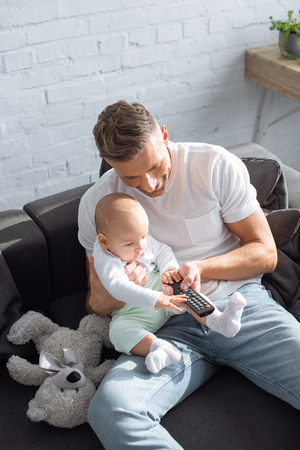 father and baby daughter sitting on couch, holding remote controller and watching television at homeの写真素材