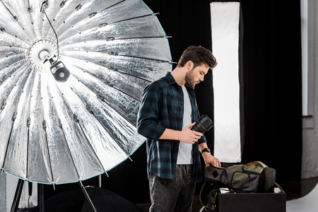 handsome young photographer holding lens and looking into backpack in photo studioの写真素材