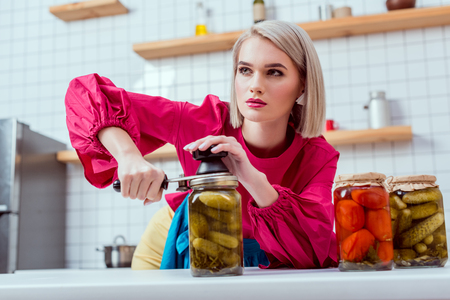 selective focus of beautiful fashionable housewife sealing jar of pickled cucumbers in kitchenの写真素材