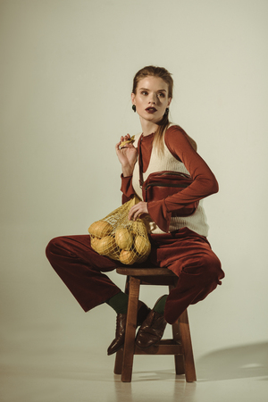 fashionable girl sitting on stool with lemons in string bag on beigeの写真素材