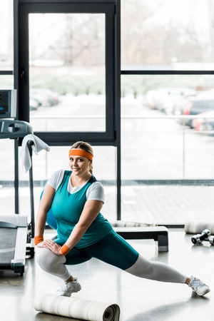 happy plus size girl in sportswear stretching near fitness matの写真素材