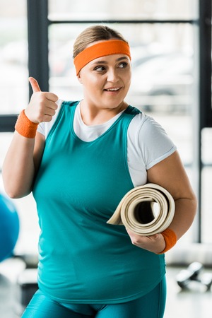 cheerful overweight woman showing thumb up while holding fitness mat in gymの写真素材