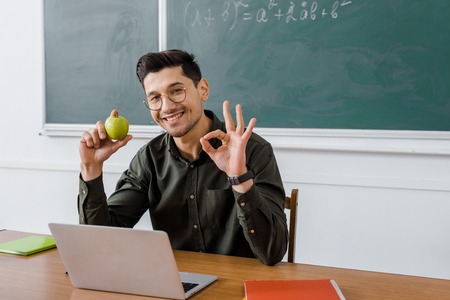 smiling male teacher in glasses holding apple and showing ok sign at computer desk in classroomの写真素材