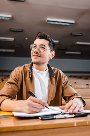 smiling male student in glasses sitting at desk and writing in notebook during lesson in classroom