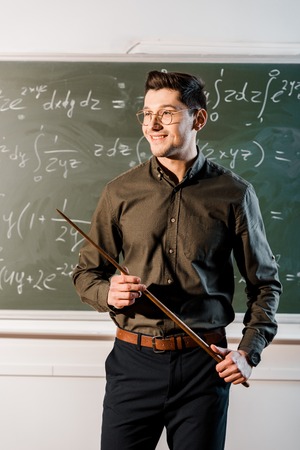 smiling male teacher in formal wear holding wooden pointer in front of chalkboard with equationsの写真素材