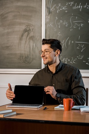 smiling male teacher in glasses sitting at desk and showing laptop with blank screen in classroomの写真素材