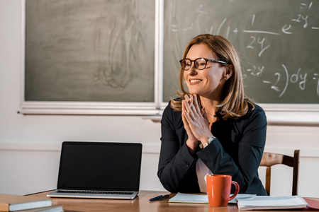 smiling female teacher sitting at computer desk with blank screen in classroomの写真素材