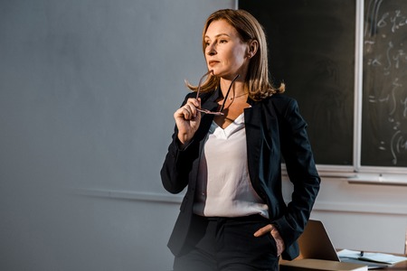 female teacher in formal wear holding glasses and looking away in classroom