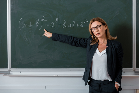 female teacher looking at camera and pointing with finger at mathematical equation on chalkboard in...