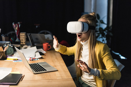 excited casual businesswoman gesturing with hands while having virtual reality experience at computer desk in officeの写真素材