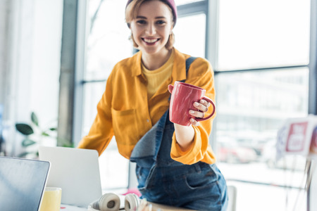smiling young female it specialist at computer desk holding cup of coffee and looking at camera in loft officeの写真素材