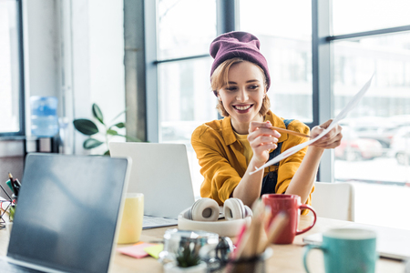 smiling young female it specialist holding document and pen while sitting at computer desk in loft officeの写真素材
