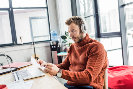 casual businessman in headphones with document sitting at computer desk in loft officeの写真素材