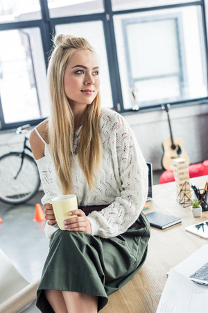 beautiful casual businesswoman holding coffee cup and looking away in loft officeの写真素材