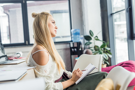 focused casual businesswoman sitting and writing in notebook in loft officeの写真素材