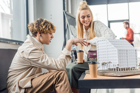 female and male architects with coffee to go working on house model in loft office