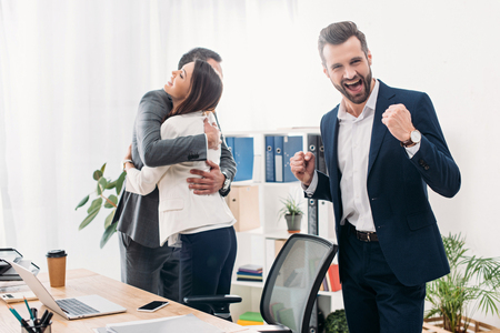 colleagues hugging, smiling and celebrating near table in officeの写真素材