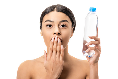 shocked young african american woman holding bottle of water and looking at camera isolated on white