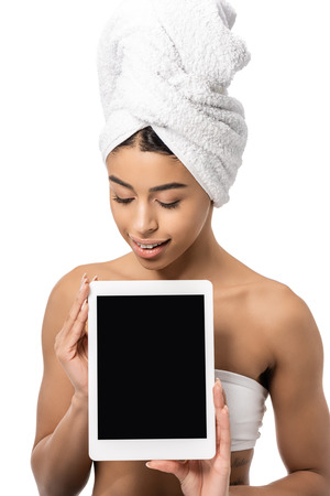 attractive smiling african american girl with towel on head looking down at digital tablet with blank screen isolated on whiteの写真素材