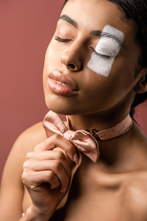 sensual african american woman with closed eyes and white paint on face adjusting bow tie isolated on brownの写真素材