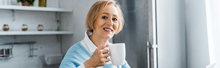 smiling senior woman holding cup of coffee and looking at camera at home