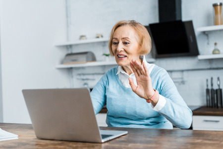 smiling senior woman sitting at table and waving with hand while having video call on laptop in kitchenの写真素材