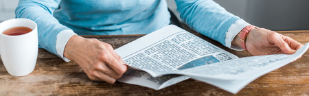 cropped view of senior woman sitting at table with cup of tea and reading newspaper at home