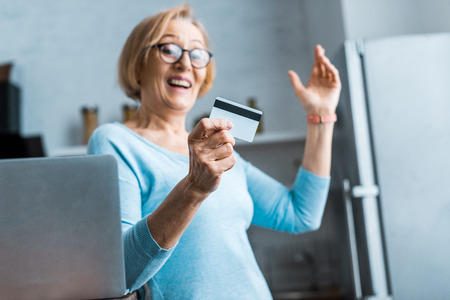 excited senior woman in glasses gesturing with hand and holding credit card near laptop at home