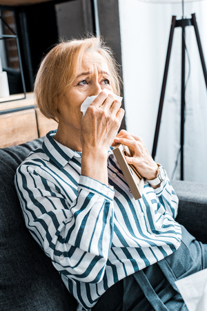 sad senior woman wiping face from tears with tissue and crying while holding picture frameの写真素材