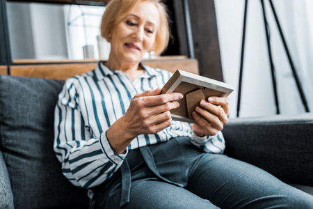 senior woman in casual clothes sitting on couch and looking at picture frameの写真素材