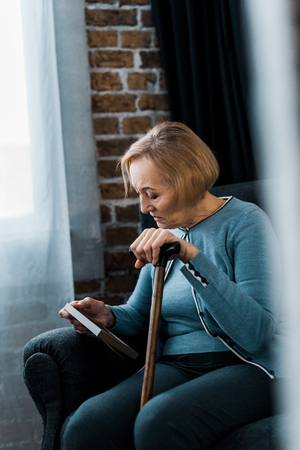 upset senior woman sitting with walking stick and looking at picture frameの写真素材
