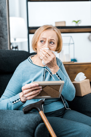 upset senior woman wiping face from tears, crying and holding picture frameの写真素材