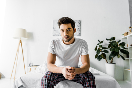 handsome serious man in pajamas sitting on bed at homeの写真素材