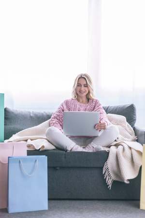 happy blonde woman sitting on sofa with laptop near shopping bagsの写真素材