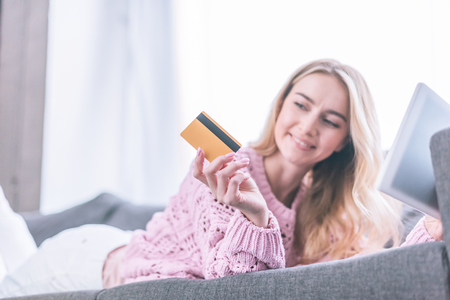 smiling woman looking at holding credit card while lying on sofa with digital tabletの写真素材