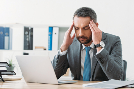 handsome businessman sitting at table with laptop and feeling headache in officeの写真素材