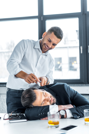cheerful man pointing with marker on face of sleeping coworker in modern officeの写真素材