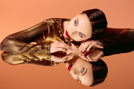 attractive woman in golden clothes with glamorous makeup and mirror reflection isolated on orangeの写真素材